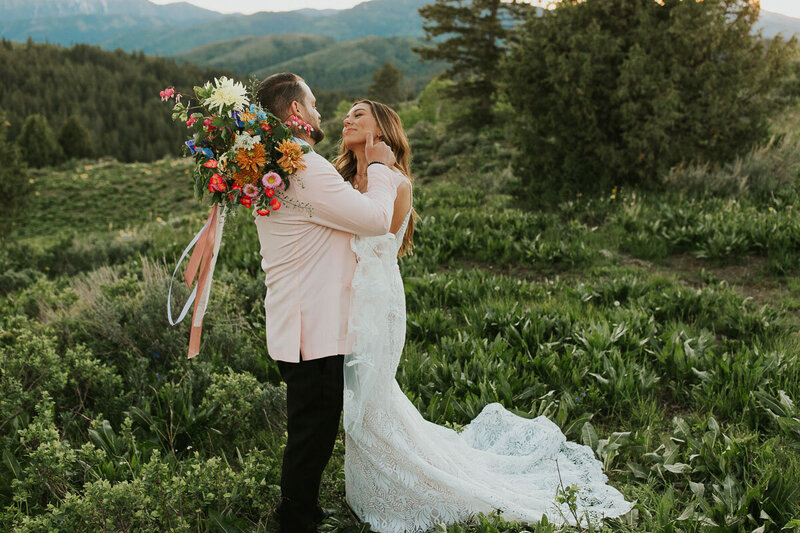 Adriana Montoya Photo, wedding and elopement photographer located in Rexburg, Idaho. A  groom is gently caressing his bride's cheek.  They are standing facing each other, so that you can see the bride's face but not the groom's. They are standing in a thick, grassy field with mountains and trees behind them.