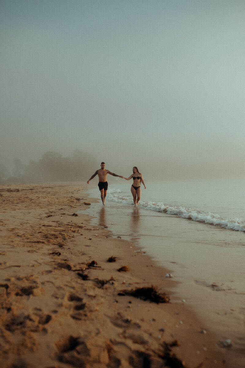 A family of three stands in shallow ocean water at the beach. The man is holding a baby, and the woman is standing next to them, both adults smiling at the child.