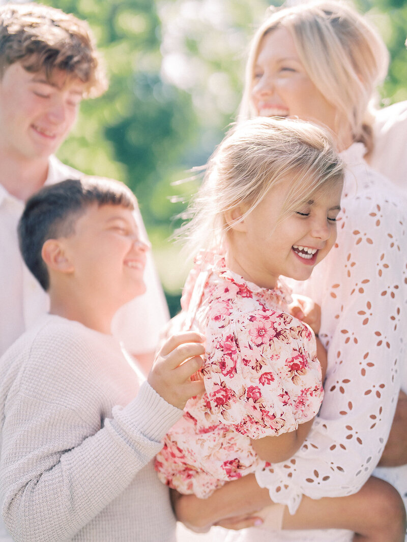 Little girl with blonde hair in pink floral dress laughs as she is tickled by her older brother and held by her mother, photographed by Northern Virginia Family Photographer Marie Elizabeth Photography.