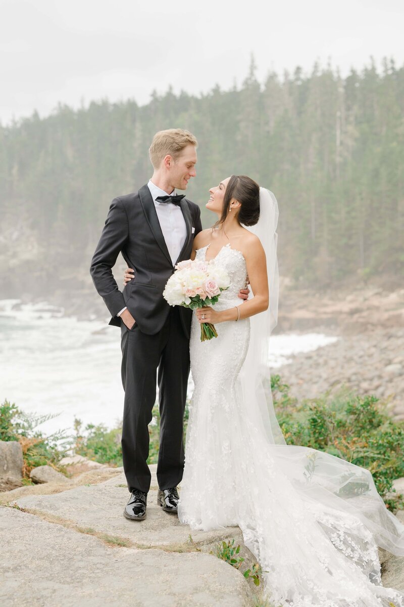 Wedding portrait of a bride and groom at Acadia National Park in Maine, standing on the rocky coastline with the ocean and forest in the background. The couple shares an intimate moment, smiling at each other while the bride holds her bouquet. Felisha, a destination wedding photographer based in New England, specializes in capturing timeless moments in Acadia National Park, Maine, New England, and worldwide locations.