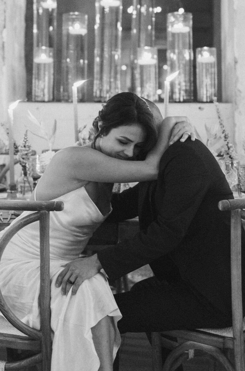 The bride and groom sit on chairs near the wedding table, leaning towards each other and embracing