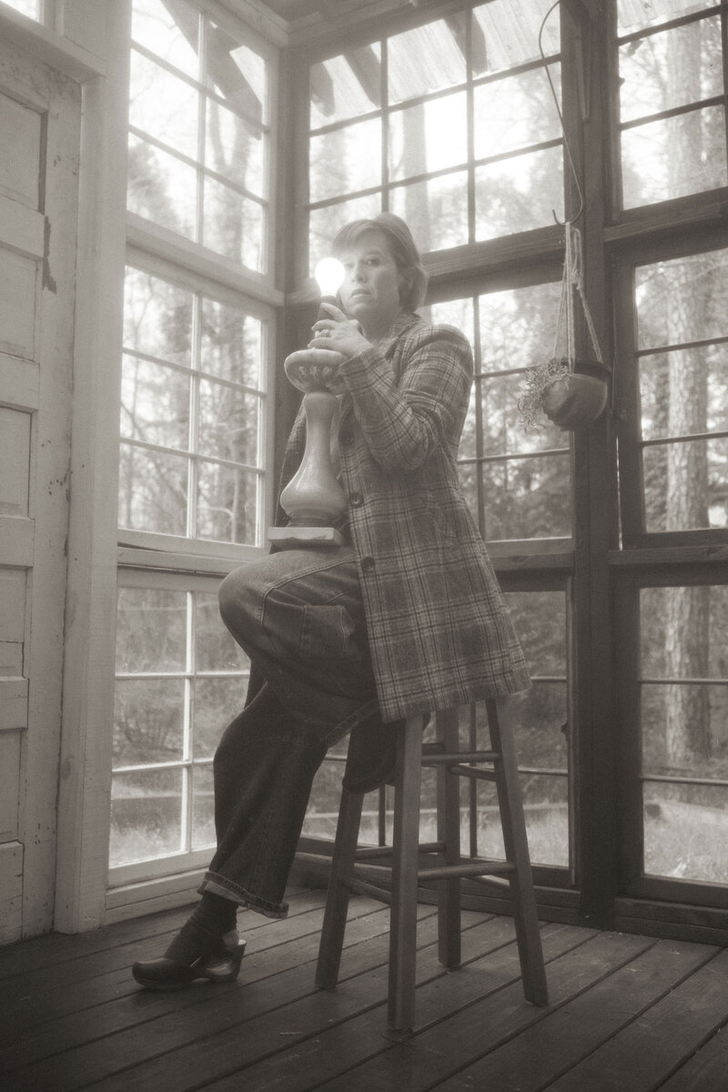 Photographer Melanie Dalton sitting on a stool at Das hinterhaus holding a shadeless lamp. 
