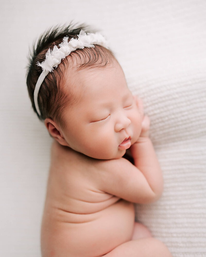 beautiful portrait of infant girl with black hair sleeping on white blanket. She is wearing a white headband and has her hand under her chin