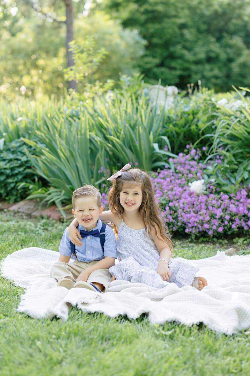 Brother and sister sitting on a white quilt in a garden during their family portraits by missy marshall