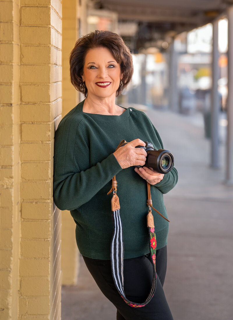 Photographer Paula Goforth holds her camera with a focused and professional demeanor. She is ready to capture a moment, embodying her passion and expertise in photography, with a background that reflects her creative environment. Paula is based out of Fort Worth, Texas serving the DFW metroplex. 