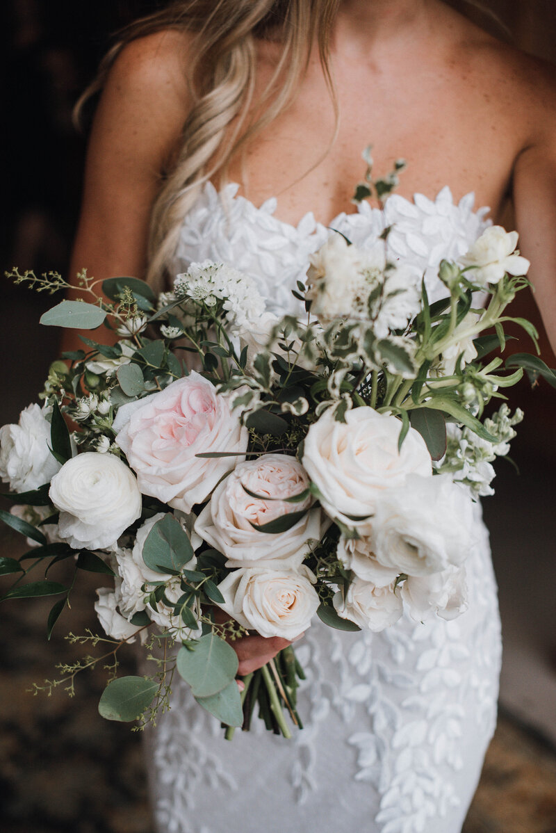 A bride in a strapless richly eembellishedd wdding gown holds a bouquet of white and blush garden roses and ranunculus