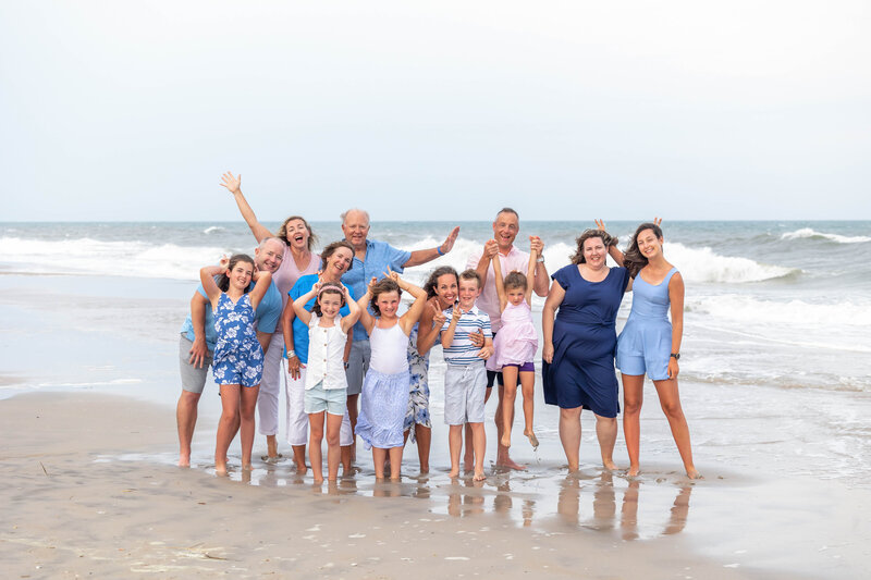 A large family joyfully poses on the shore of Ocean Isle Beach, with children and adults dressed in blue and white outfits. The ocean waves provide a lively backdrop, highlighting the fun of a family day at the beach. Ideal for artistic family photography in Wilmington, NC.