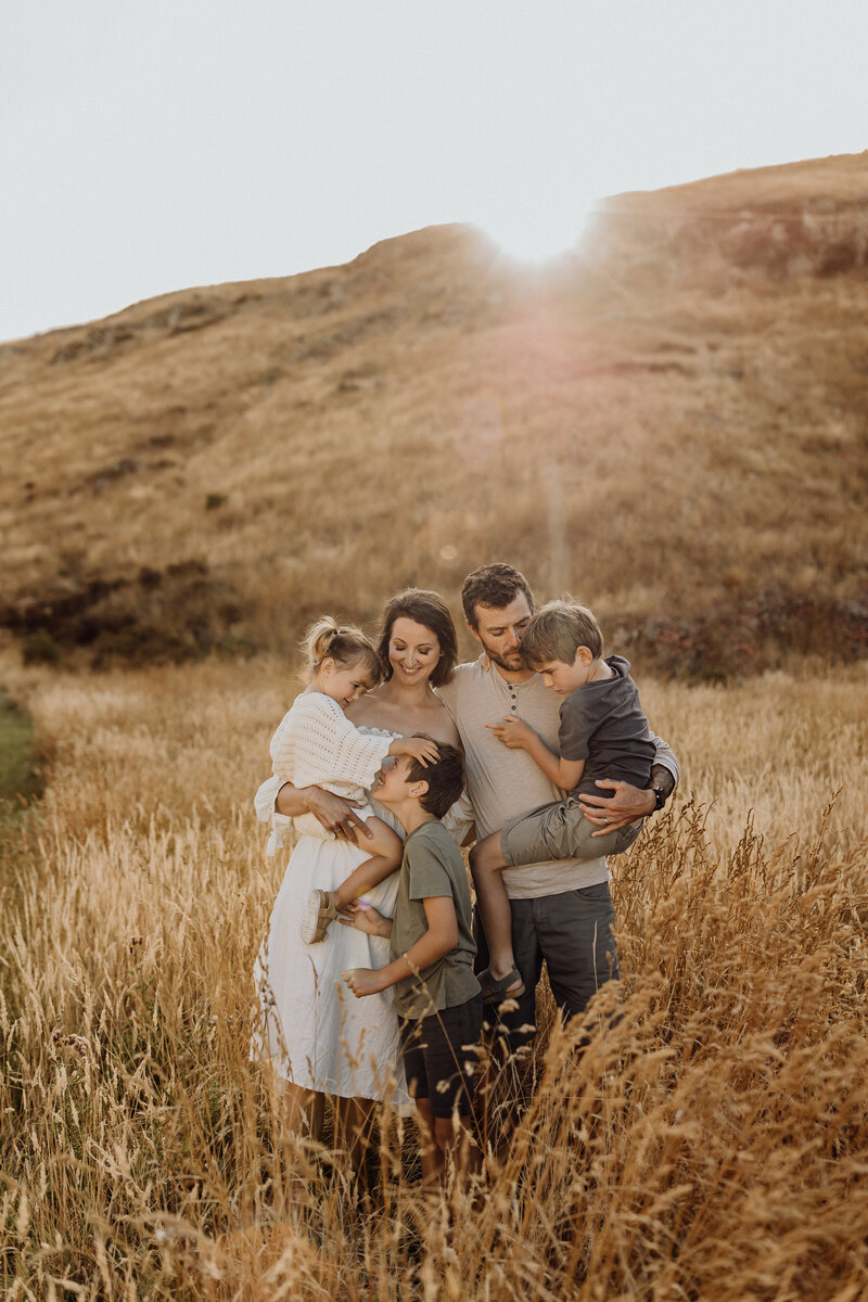 A family of five holding one another while photographed by Christchurch photographer Lucy Jane Photography in a field of long grass at sunset