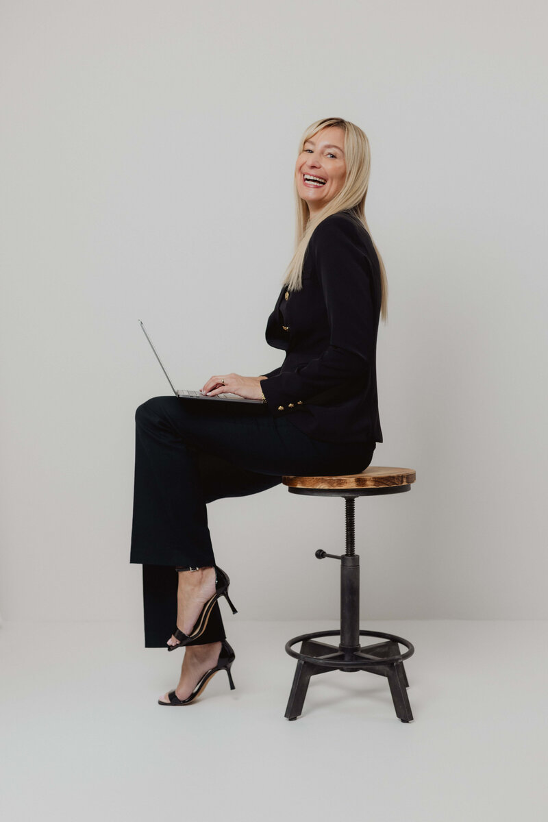 A omen in a black suite on a lap top.  She is smiling and appearing confident.