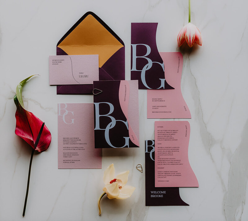 Unique and vibrant wedding invitations in a pink and purple colour palette and bold white text. Invitations paired with a purple and dark yellow envelope,  and matching wedding menu and escort card.