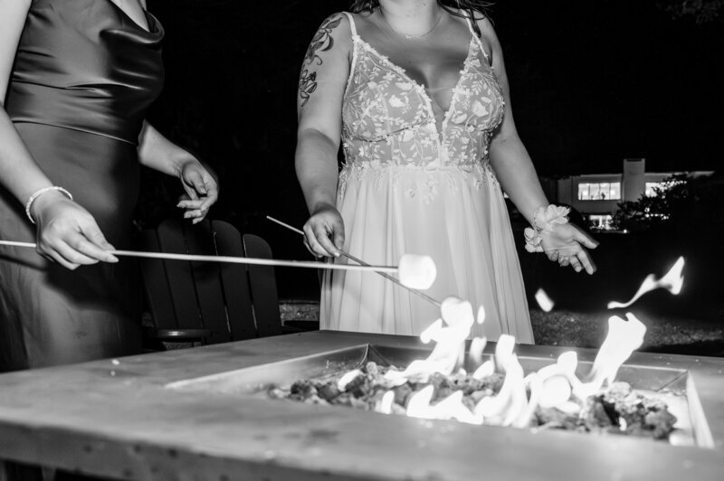 black and white of curvy bride in spaghetti strap gown holding a marshmallow on a metal stick over a fire pit 