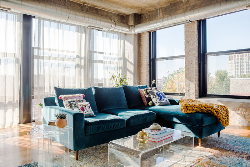 The bright and airy living room of The City Loft by Indigo Ink Designs. A plush teal velvet sectional sofa with colorful patterned pillows and a chunky mustard yellow knit blanket sits on a patterned rug. A clear acrylic coffee table holds books and a gold decorative object. Sunlight streams through large windows covered with sheer white curtains, highlighting the exposed brick wall and industrial ductwork on the ceiling. A small clear acrylic side table holds a potted plant.