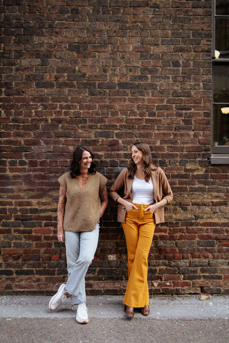 Two women stand smiling and tlaking in front of a brick wall. One wars a brown top and light jeans with white sneakers, the other wears a brown jacket, white top, yellow pants, and brown shoes. 