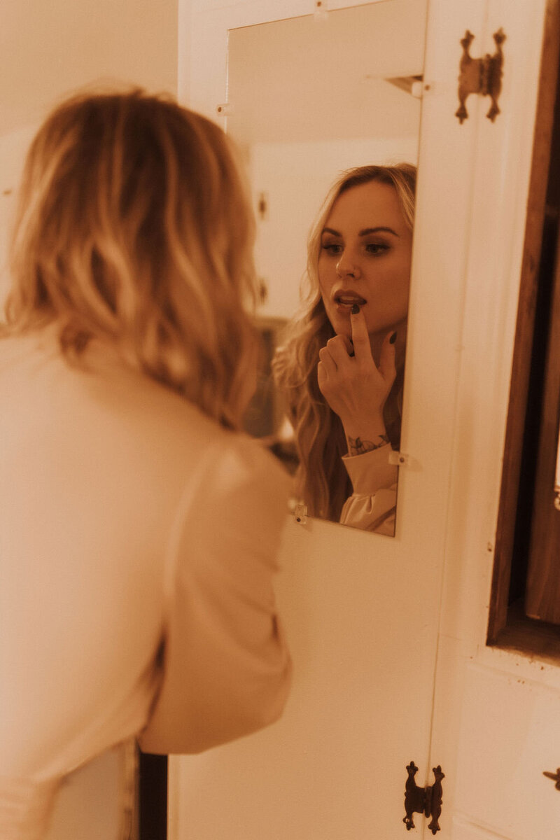 Hair extension professional is looking in the mirror and applying lipstick. She is wearing an elegant white blouse.