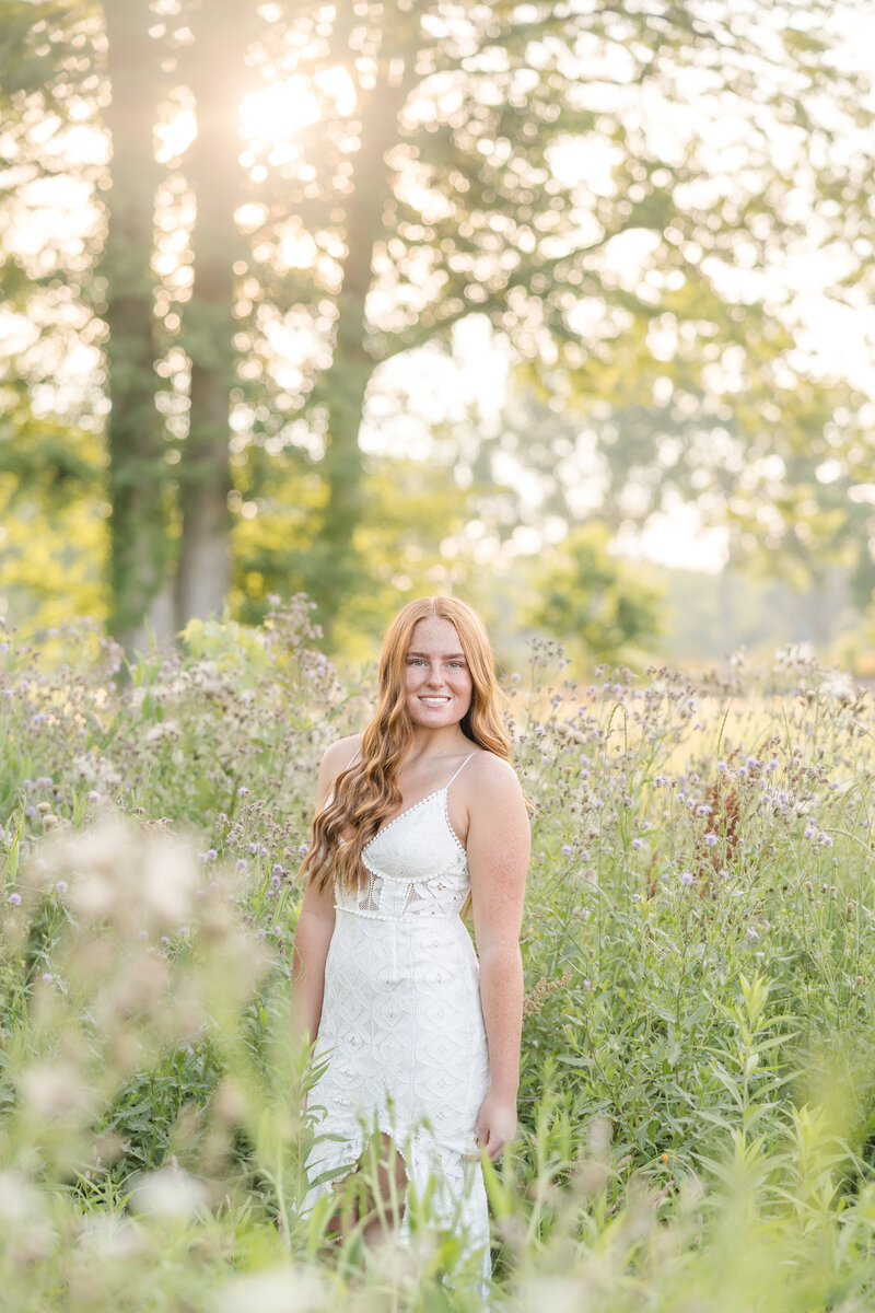 A red haired senior in a white dress standing in a field of wild flowers at golden hour during a senior portrait session. 