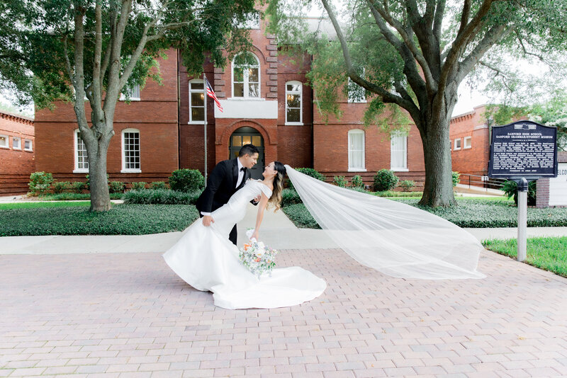 Bride and groom share a joyful moment outside Venue 1902; groom looks at bride as she smiles holding a pastel bouquet, wearing a long veil and satin gown under a large oak tree.