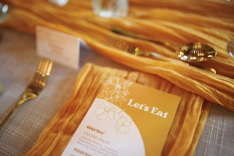 A retro mustard wedding menu sit on a mustard yellow cheesecloth napkin at table runner at a wedding at Evermore in Almonte, designed by Frid Events.