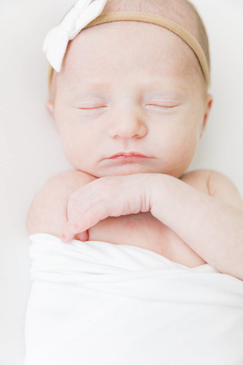 A newborn baby girl wearing a white bow sleeps with a hand under her chin
