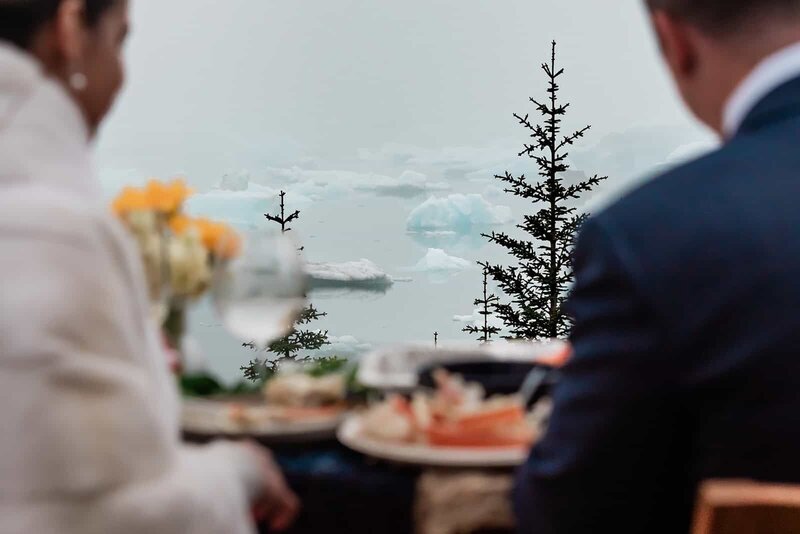 A bride and groom are slightly out of focus on the edge of the frame sitting at their wedding dinner in their wedding clothes--between them in clear view are the large blue-tinted white icebergs floating in Bear Lake during their two day luxury elopement with dogsledding in Alaska.  On the table you can see hint of orange and tan piles of Alaskan crab legs, a wine glass, and a pop of yellow flowers.  