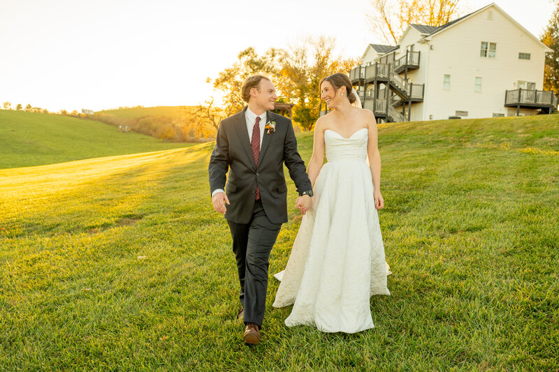 Bride and groom walking hand in hand on a grassy hill during golden hour, smiling at each other. Wedding at Batten Green in Staunton, VA. Captured by Mason Jesmer Photography, Richmond wedding photographer.