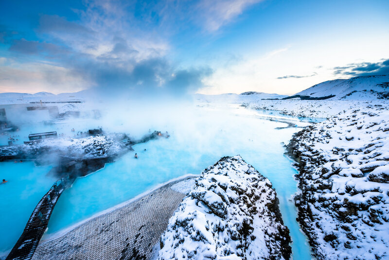 Scenic view of a geothermal lagoon surrounded by snow-covered rocks and hills under a blue sky. Steam rises from the turquoise water, contrasting with the white landscape. Visitors are visible enjoying the hot springs.