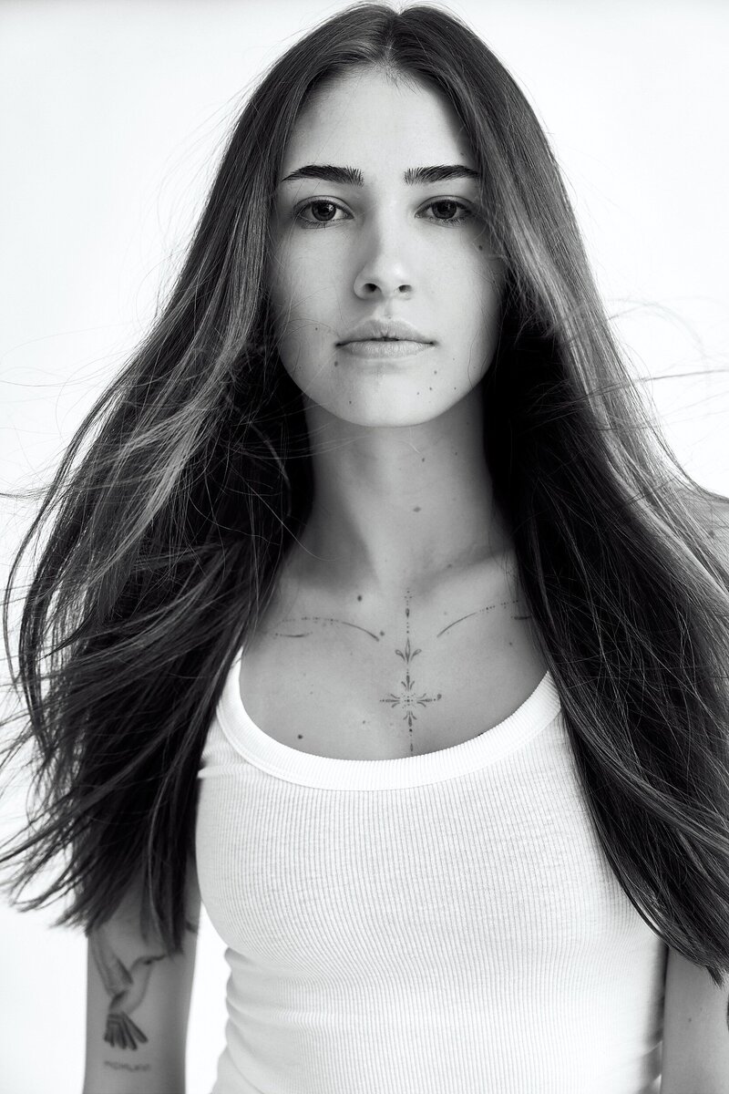 Black and white portrait of a young woman with long flowing hair and minimal makeup, wearing a ribbed white tank top. She has visible tattoos on her collarbone and arm, standing against a clean white background.