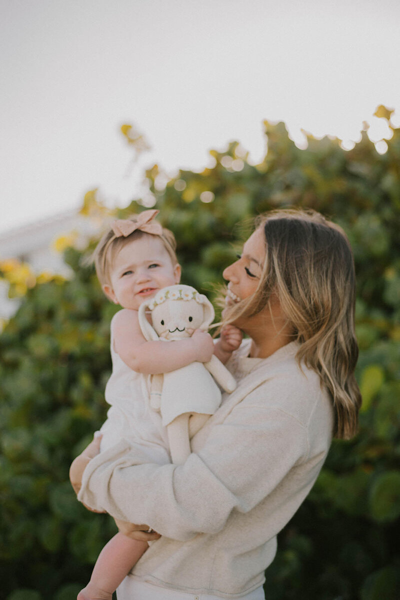 Baby sleep consultant smiles against stone wall in tan sweater and white jeans