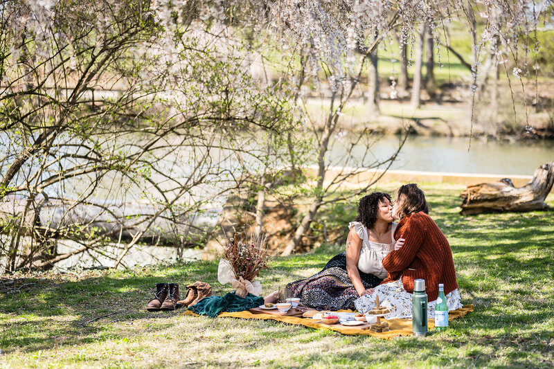 An LGBTQ+ couple sit on a picnic blanket with various foods and drinks sprawled across. The pair leans into one another for a kiss under the shady trees at Duck Pond.