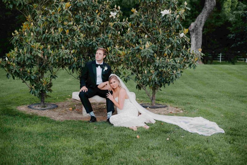 A bridal couple poses outdoors for a photo with the groom sitting on a stone bench under a blooming tree and the bride seated on the grass next to him, her veil fanned out behind her, both looking towards the camera.