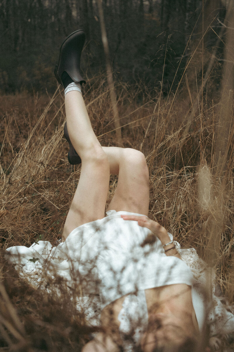 A pregnant woman in a white dress laying in a field of straw kicking her legs up in the air. 