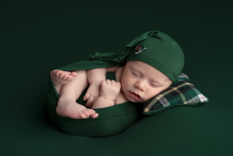 Cozy newborn portrait of a baby boy in a wrapped Huck Finn pose with plaid pillow, styled in rich greens – Paris, Ontario.
