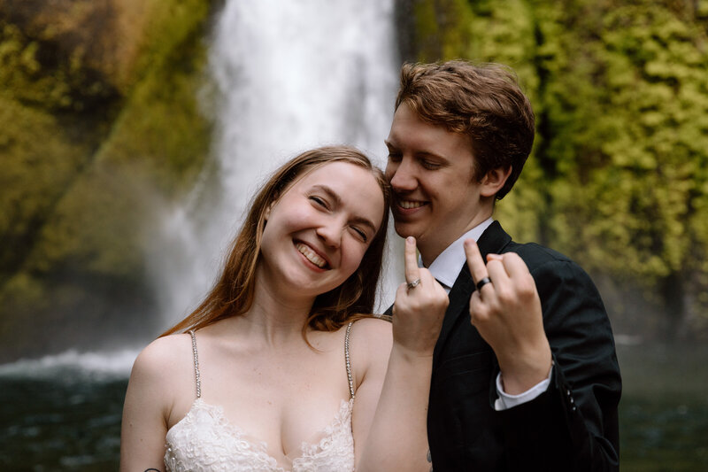 Couple holding out their ring fingers while standing in front of a waterfall in the Columbia River Gorge.