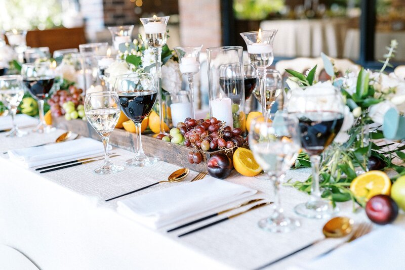 Elegant wedding table setting with gold flatware, wine glasses, fresh fruit, white flowers, and candles arranged in a wooden centerpiece box.