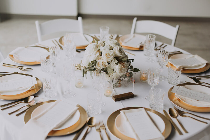 A wedding table with lush white rose centrepieces, gold chargers, and crystal goblets set by the window at Mainspace Canmore.