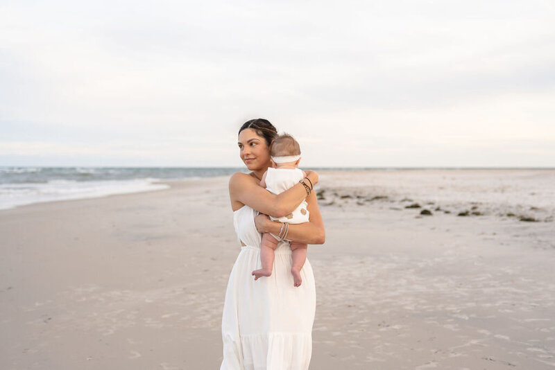 mother holds her baby with it's back facing the camera as she gazes over the ocean on a gorgeous, clear day