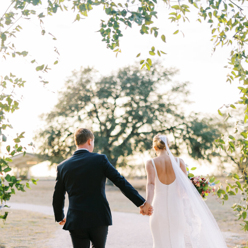 Bride and groom holding hands walking away from the camera with trees in the background