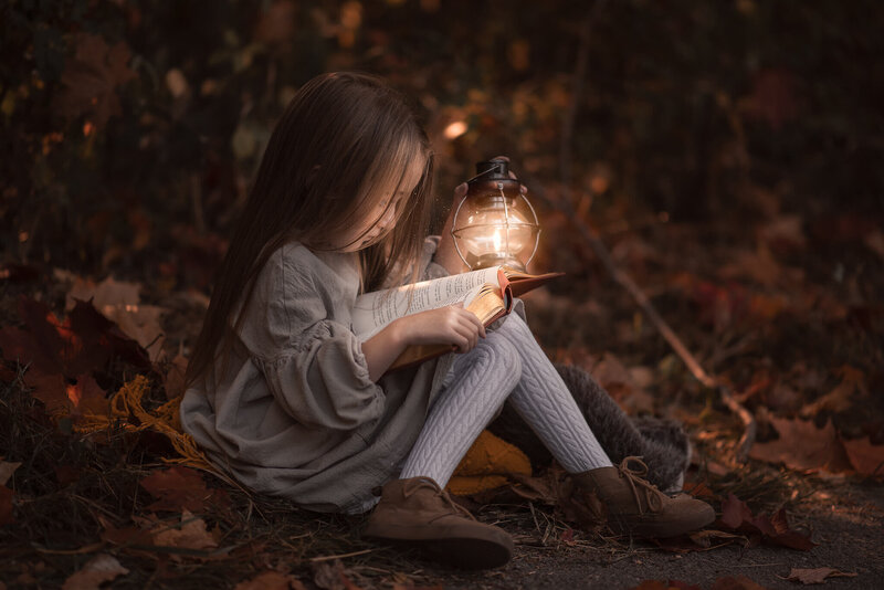 Captured in Kamloops by Brittany Danielle is a portrait of a little girl with long brown hair sitting on the ground. She is holding a glowing lantern up on her knees and looking into it.