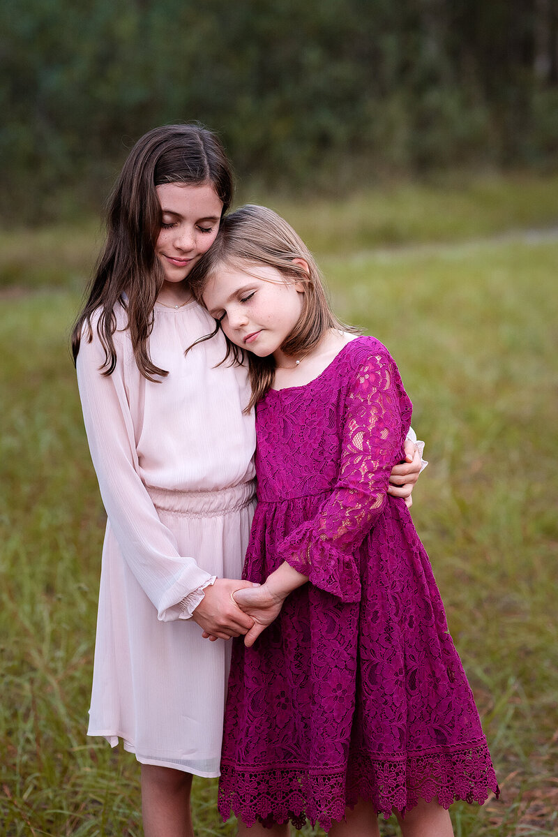 Sisters dressed in pink and fuchsia dresses snuggled together with greenery background at Julington Creek Preserve in FL.