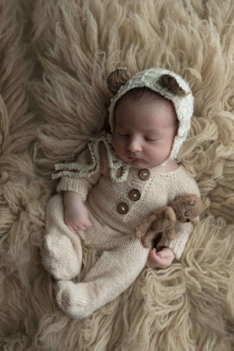 Creamy textures and whimsical bear details surround this baby boy, captured sleeping in a soft knit romper and bonnet on a flokati rug.