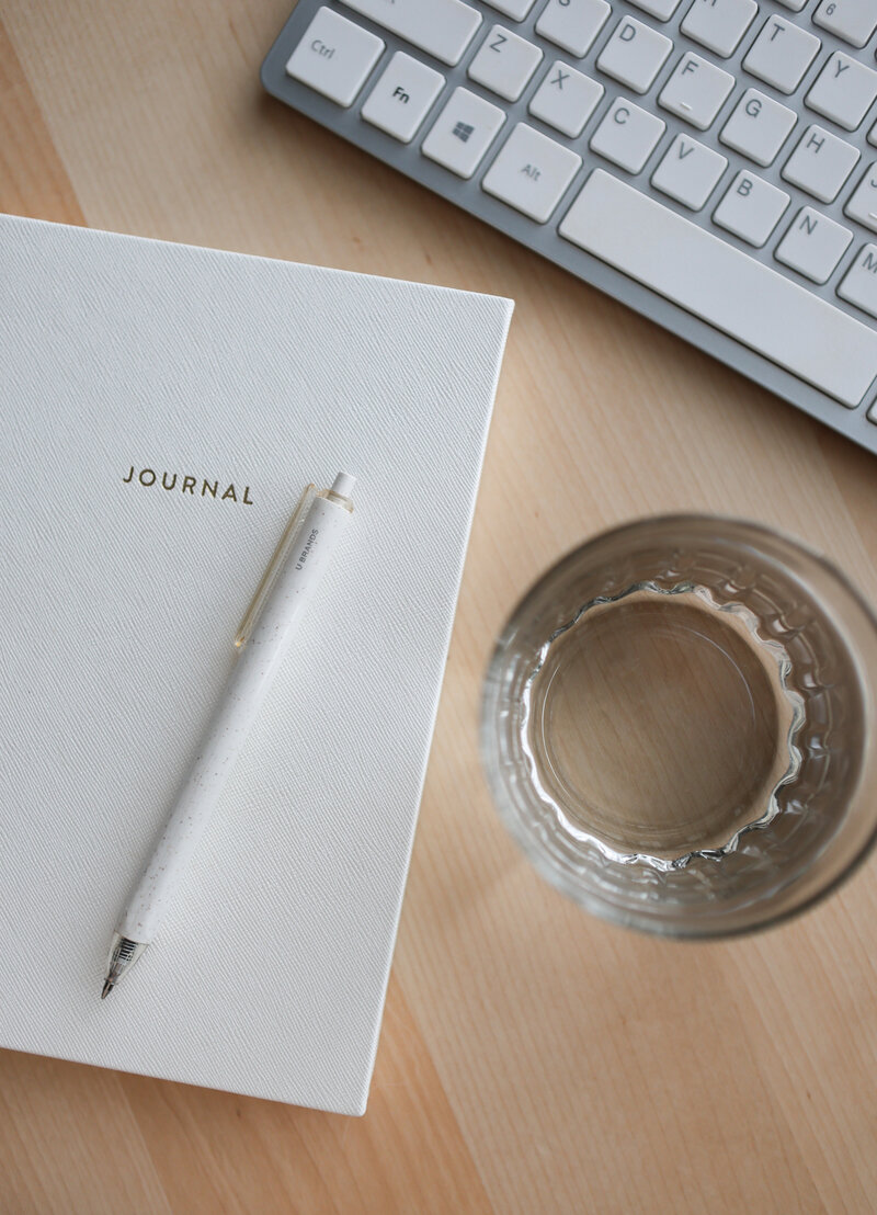 Photo of Apple keyboard, white journal, glass of water and pen