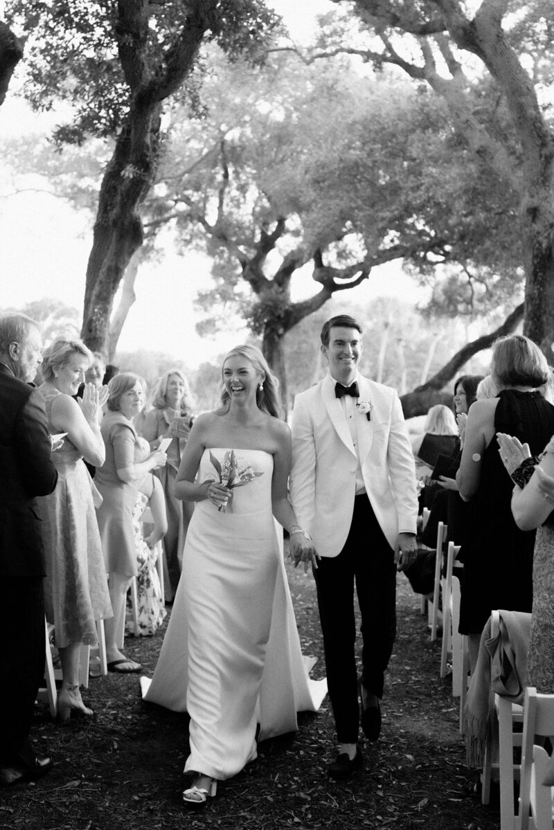 Bride and groom walking down an outdoor aisle together while guests applaud.