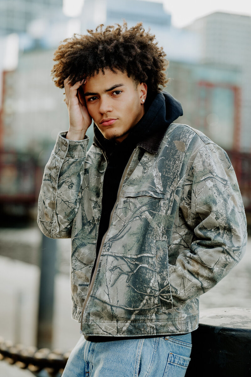 Boy with the city of Boston behind him  wearing a camo jacket with his hand in his pocket 