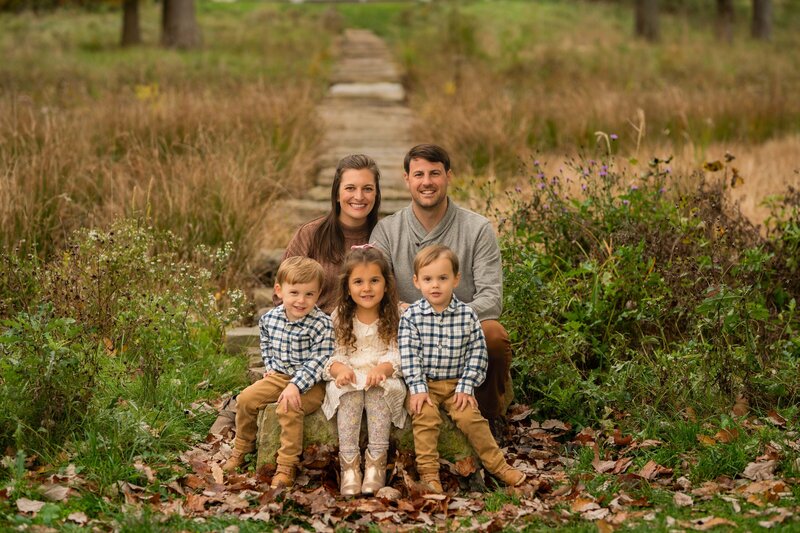 mom and dad sitting on a rock with their triplets all sminig at the camera on a fall day in cleveland ohio