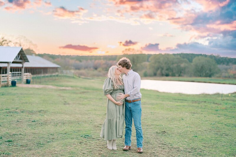 Pregnant woman in green dress kisses husband in field with a pink and orange sunset