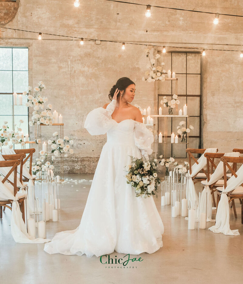 Bride in off-the-shoulder gown posing in front of ceremony aisle lined with Crossback chairs, white florals, and candles.