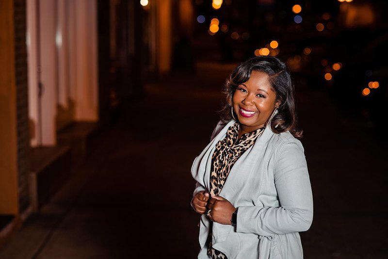 Night photo of small business owner standing outside wearing cute leopard print top and dusty blue jacket, smiling at the camera with lights behind her