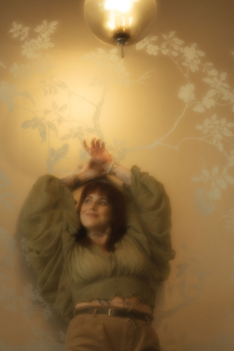A woman with tattoos poses against a gold wallpapered wall.