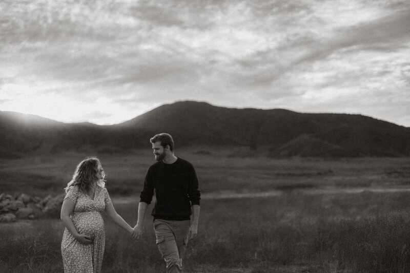 Expectant couple walking hand-in-hand through a wide open field, captured in black and white at golden hour in Menifee, CA.