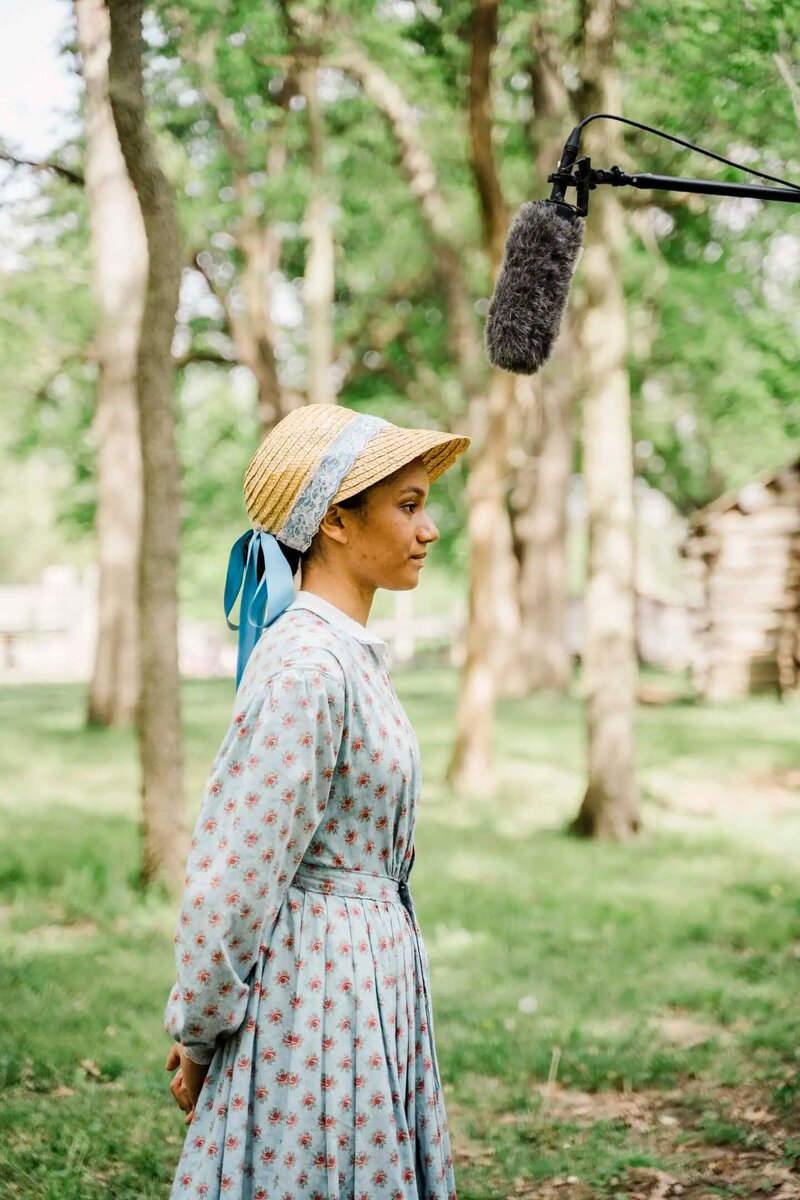Young woman in 19th-century period costume stands outdoors in a wooded area, facing a boom microphone during a historical film production.