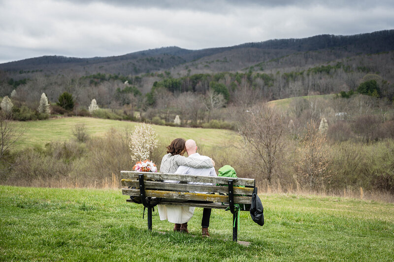 A couple on their elopement day sit on a park bench on top of a hill with their hiking backpacks. The couple wrap their arms around one another and face the view of the Blue Ridge Mountains from Heritage Park in Blacksburg, Virginia.